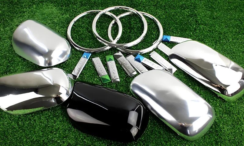 Assorted chrome and black car accessories arranged on grass, including mirror covers and trim rings