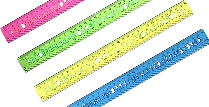 Four colorful transparent plastic rulers in pink, green, yellow, and blue, each marked with both centimeter and inch measurements and alphabet cutouts