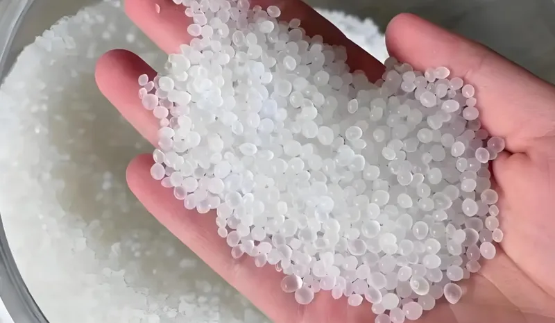A hand holding translucent plastic pellets above a container of more pellets